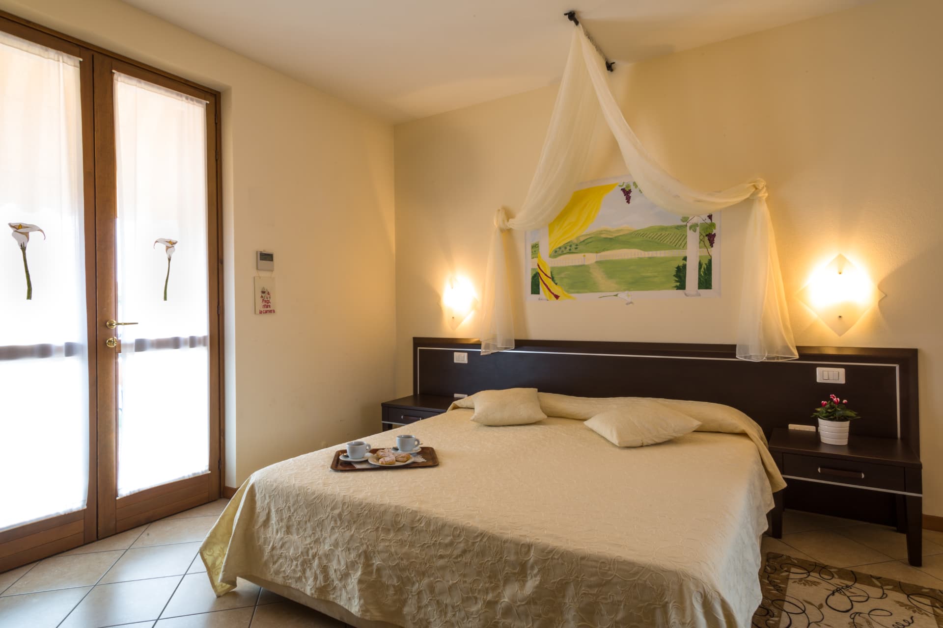 Camera delle calle: letto con copriletto crema e baldacchino bianco sotto l'affresco di colline con calle